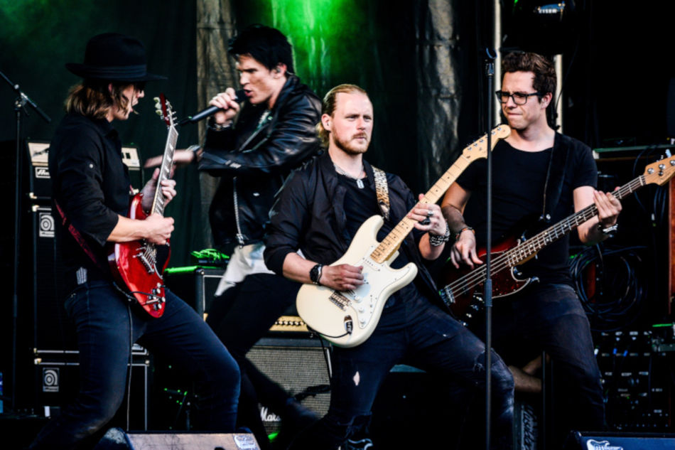 Four men rock band. One on electric guitar in front, and one on vocals, one on electric guitar, 
                            one on bass in background
